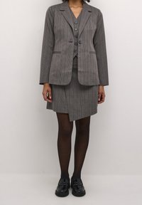 Šedý kostkovaný oblek skládající se z přizpůsobeného saku s klopovým límcem a odpovídající sukně ke kolenům. Zdobený černými knoflíky.