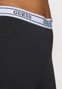 Crni pamučni bokserice s bijelom elastičnom trakom oko struka koja sadrži "GUESS" logo u crnom tekstu i dvojne crne pruge duž vrha.
