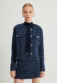 Marineblau gestreifte Jacke mit metallischen Akzenten und zwei Brusttaschen, kombiniert mit einem passenden Rock mit Faltenwurf.