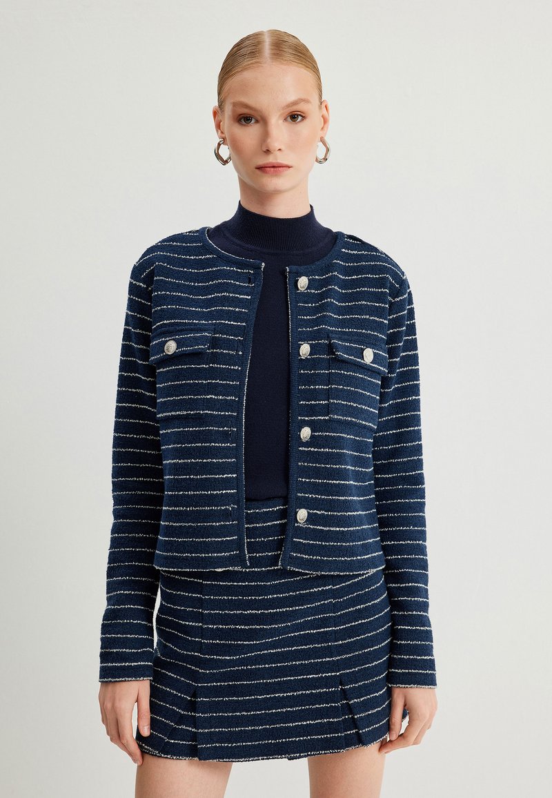 Marineblau gestreifte Jacke mit metallischen Akzenten und zwei Brusttaschen, kombiniert mit einem passenden Rock mit Faltenwurf.