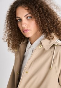 Beige jas met gouden accenten, met een ritssluiting en een trekkoordcapuchon. Witte overhemd met kraag eronder, met een gladde textuur.