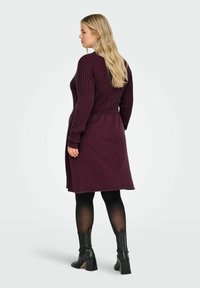 Adīta kleita dziļi burgundijas krāsā ar ribotu tekstūru, garām piedurknēm un sašaurinātu jostasvietu. Valkāta ar melniem augstpapēžu zābakiem un necaurspīdīgām zeķbiksēm.