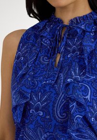 Blusa blu con motivo paisley, caratterizzata da scollo arricciato con chiusura a fiocco e dettaglio a occhiello, realizzata in tessuto strutturato.