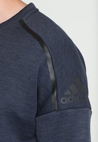 Gros plan sur une manche bleu marine Adidas avec une bande noire allant du col à l'épaule et le logo Adidas près du poignet.