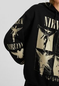 Sudadera con capucha negra de corte extragrande, con estampados gráficos dorados de figuras angelicales y la palabra "NIRVANA" en letras llamativas. Capucha ajustable con cordón.
