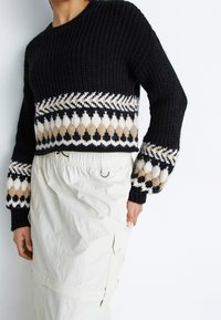 Czarny sweter dziany z wzorzystą beżowo-białą ozdobną wstawką; posiada szerokie rękawy i okrągły dekolt. Połączony z białymi spodniami.