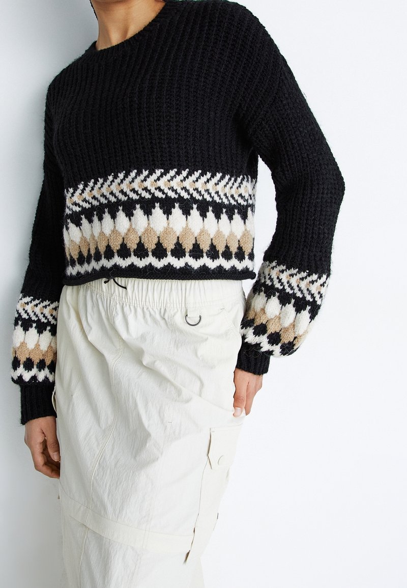 Czarny sweter dziany z wzorzystą beżowo-białą ozdobną wstawką; posiada szerokie rękawy i okrągły dekolt. Połączony z białymi spodniami.