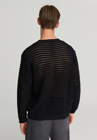 Uomo che indossa un maglione nero a maglia aperta che mostra la pelle sottostante, abbinato a pantaloni grigi, in piedi contro uno sfondo chiaro e neutro, voltato di spalle.