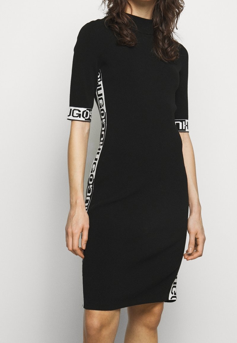 Femme portant une robe noire longueur genou avec des manches courtes et des panneaux latéraux blancs affichant un texte noir audacieux.