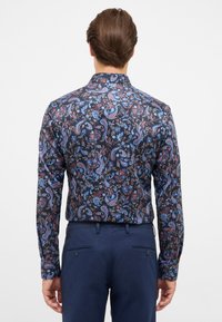 Langsleeve shirt in donkere stof met een multikleurig bloemen- en paisleypatroon. Effen marineblauwe broek met achterzakken.