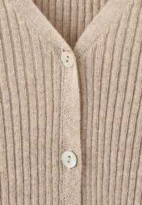 Detailní záběr na béžovou žebrovanou pleteninu se dvěma bílými knoflíky zapnutými podél svislého středového lemu.