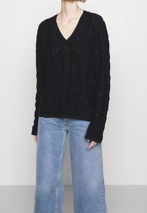 Femme portant un pull en V texturé noir et un pantalon large en denim bleu clair, se tenant contre un fond gris clair uni.