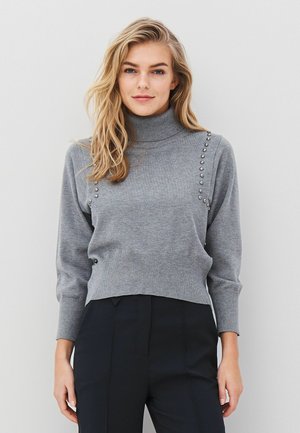 Grå polotröja i mjuk stickad tyg, med dekorativa metallnaglar längs den vänstra axeln och en kortad fåll.