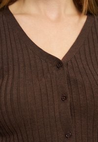 Κοντινό πλάνο ενός ατόμου που φορεί μια καφέ ριγωτή ζακέτα με τρία ορατά κουμπιά και V-λαιμόκοψη.