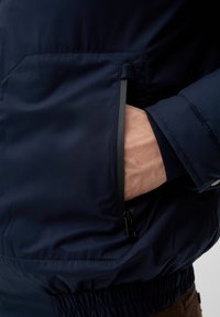 Mørkeblå jakke med en lynlåslomme. Har elastisk talje og håndled. Glat stof med subtil glans, der fremhæver minimalistisk design.