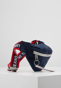 Borsa a vita in nylon blu navy con forma triangolare, zip argentata, tracolla regolabile rossa e bianca, e un'etichetta a forma di portachiavi attaccata.