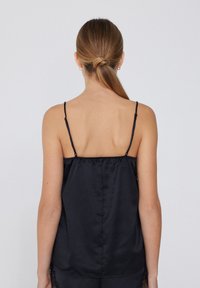 Camisole en satin noir avec de fines bretelles réglables, présentant une texture lisse et une bordure en dentelle le long de l'ourlet. Vue de dos.