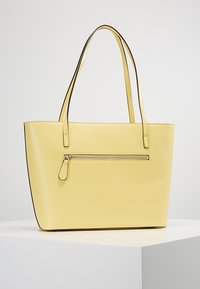 Żółta torba typu tote ze sztucznej skóry z dwoma paskami na ramię, kieszenią na zamek z przodu oraz prostokątnym, usztywnionym kształtem. Prosty wzór bez nadruków.