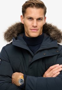 Marineblaue Winterparka mit abnehmbarem, fellgefüttertem Kapuze, Reißverschluss vorne und aufgesetzten Taschen. Getragen über einem gerippten Strickrollkragen.