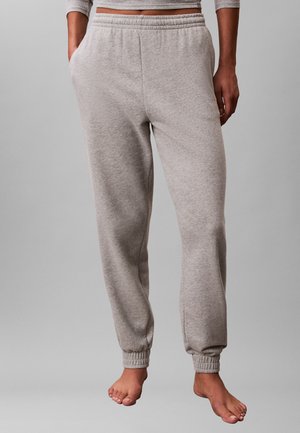 Personne portant un pantalon de jogging gris clair avec une taille et des poignets élastiques, debout pieds nus devant un fond gris uni.