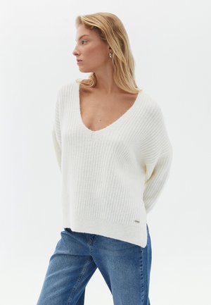 White ribbed knit sweater with a V-neck, relaxed fit, and dropped shoulders. Soft texture and side slit, paired with blue jeans.