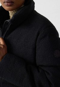 Gros plan sur une personne portant une veste noire texturée par-dessus un pull col roulé beige, tenant le col de la veste d'une main.