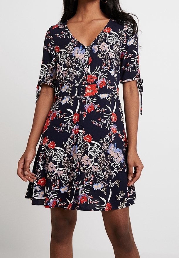 Femme portant une robe fleurie bleu marine avec des fleurs rouges, blanches et violettes claires, des manches courtes avec des liens et un col en V.