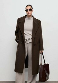 Bruine wollen overjas met grote revers, dubbele knopen en zijzakken over een lichtbeige trui en bijpassende wijde broek. Bordeauxrode handtas.