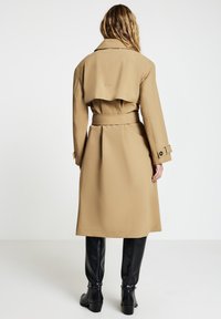 Beiger Trenchcoat mit Taillengürtel, mit Rückentasche und verstellbaren Manschetten, kombiniert mit schwarzen knielangen Stiefeln. Glatte Textur.