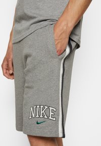 Hombre con pantalones cortos grises de Nike con franjas laterales blancas y negras, mano en el bolsillo y venas del brazo visibles sobre un fondo blanco.