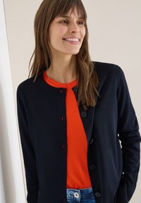 Femme souriante aux longs cheveux bruns, portant un cardigan bleu marine sur un t-shirt orange vif et un jean bleu, debout devant un fond clair.