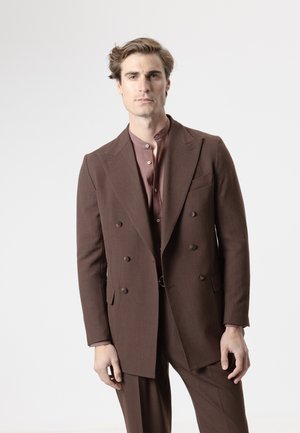 Hombre con traje marrón cruzado y camisa burdeos abotonada, de pie contra un fondo liso gris claro.