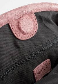 Interior de una bolsa de tela negra con una solapa de ante rosa y un botón, que cuenta con un zipper y una pequeña etiqueta. Superficies texturizadas y colores contrastantes.