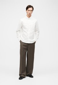 Homme debout portant une chemise blanche à boutons, un pantalon large marron et des chaussures noires, les mains dans les poches, sur un fond blanc uni.