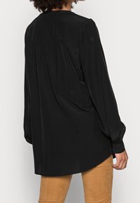 Camicia nera a maniche lunghe con bottoni, dal tessuto morbido e liscio, caratterizzata da un dettaglio arricciato sul retro e un orlo arrotondato, abbinata a pantaloni beige chiaro.
