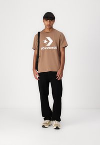 Ruskea lyhythihainen T-paita, jossa on suuri valkoinen "CONVERSE"-logo ja tähti. Yhdistetty mustiin housuihin ja vaaleisiin lenkkikengiin. Yksinkertainen muotoilu, rento istuvuus.