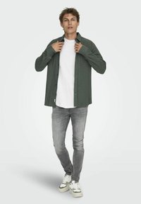 Camicia button-up verde scuro sopra una maglietta bianca, abbinata a jeans grigi chiari e sneakers bianche. La camicia presenta maniche lunghe e una vestibilità rilassata.