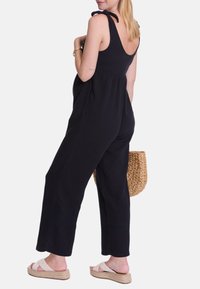 Schwarzer ärmelloser Jumpsuit mit gebundenen Trägern, lockere Passform, weite Beine, strukturiertes Material, kombiniert mit einer geflochtenen Tasche und Espadrilles.
