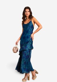 Abito maxi in satin, svasato, di colore blu scuro con un motivo floreale, presenta un orlo arricciato e sottili spalline a spaghetti. Abbinato a sandali con tacco dorati.