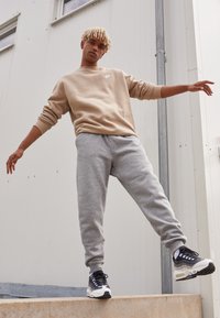 Brązowa bluza z logo Nike, szare dresy i czarno-białe sneakersy. Osoba pozuje z wyciągniętymi ramionami w miejskim otoczeniu.