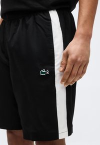 Μαύρο σορτς με λευκή κατακόρυφη ρίγα και πράσινο λογότυπο κροκόδειλου στο αριστερό μηρό, φορεμένο από άτομο με το δεξί χέρι χαλαρά στο πλάι.