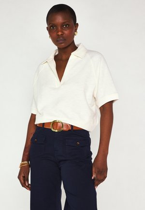Femme portant une chemise blanche à manches courtes avec col, un pantalon bleu marine foncé avec poches avant, une ceinture marron à boucle dorée et des boucles d'oreilles anneaux dorées.