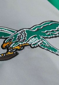 Grauer Stoff mit einem bestickten Vogel in Türkis und Weiß, mit gelben Akzenten. Das Design umfasst detaillierte Flügel und ein rundes Objekt darunter.