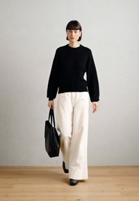 Black textured knit sweater, wide ivory trousers, and black tote bag. Model walks on wooden floor against gray background.