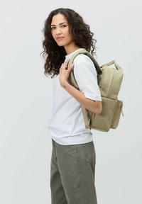 Kaki ryggsäck med flera fack, stoppade remmar och ett handtag på toppen. Bärs på en axel mot en enfärgad bakgrund.