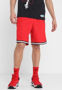 Červené sportovní šortky s černo-bílým lemováním, vyrobené z materiálu odvádějícího vlhkost. Kombinovány s červenými ponožkami a červeno-černými teniskami.