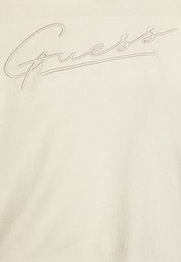 Cream sweater featuring an embroidered "Guess" logo in a wavy script. Soft, smooth texture with a fitted design and ribbed hem.