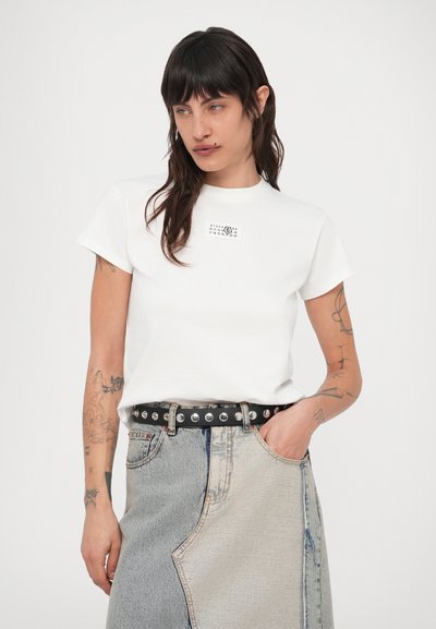 Sötét hajú, tetovált nő fehér pólót visel, amelyen kis naptár van nyomtatva, és kétrészes farmer szoknyát fekete szegecses övvel.