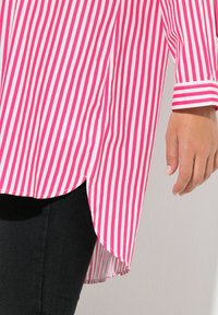 Roze-wit gestreepte blouse met een afgeronde zoom, voorzien van lange mouwen en een knoopmanchet. Zwarte broek zichtbaar onder de blouse.