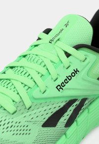 Zaļa sieta skriešanas apava ar melniem akcentiem, ar teksturētu augšdaļu, izturīgām šņorēm un polsterētu mēli, uz kuras redzams Reebok logo.